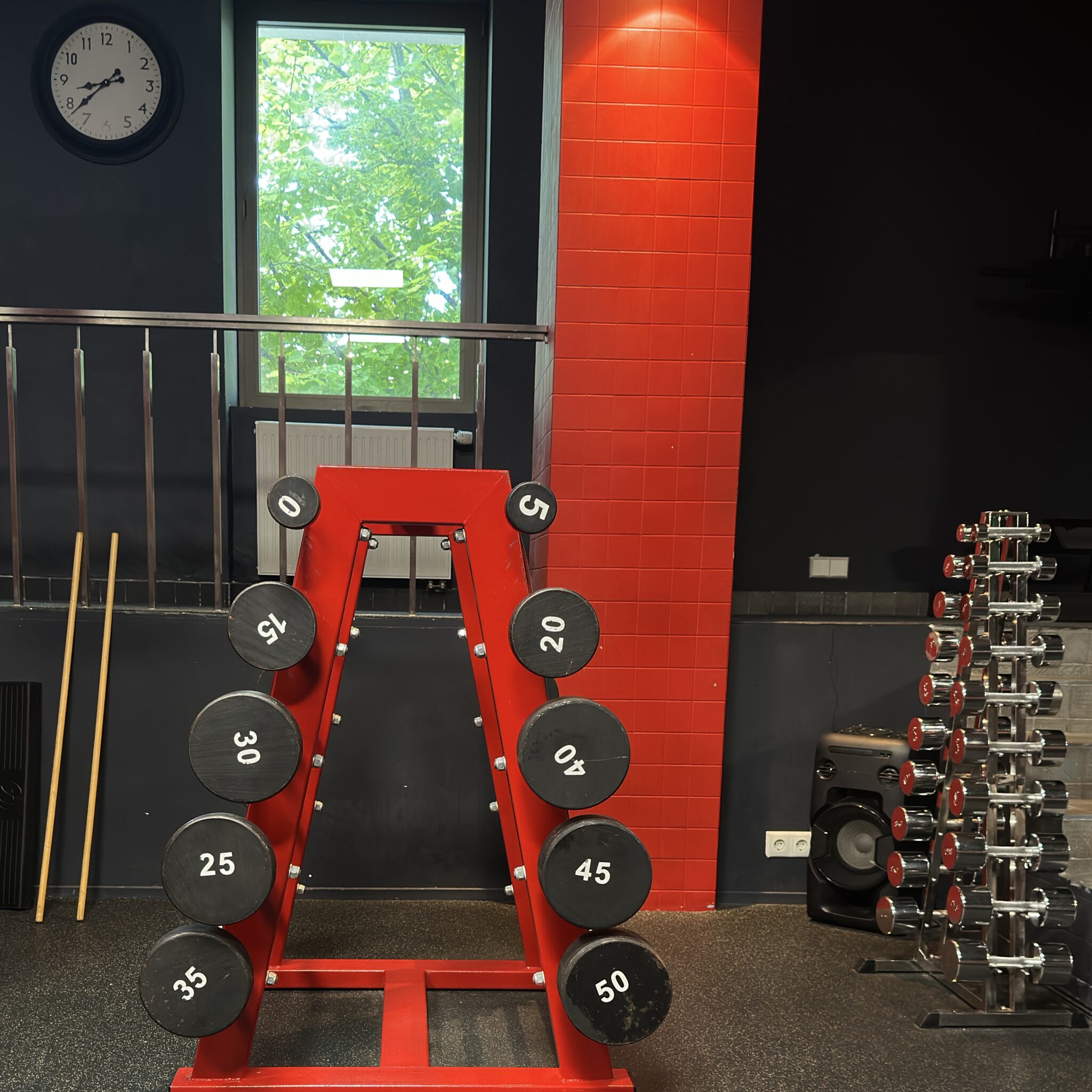 Punane hantliraam spordisaalis, taustal kell ja aken.