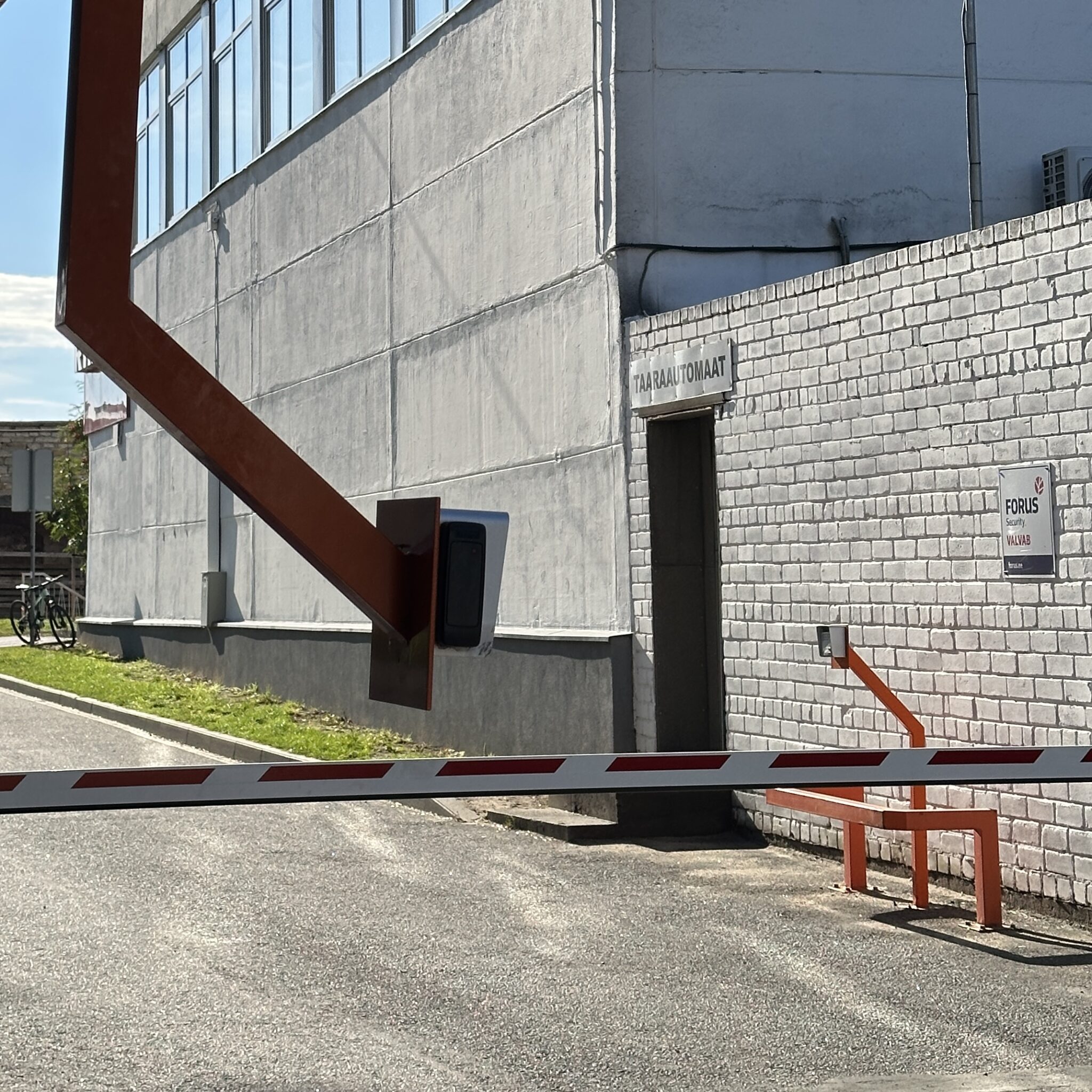 Punavalge tõkkepuu ja silt „TAARA AUTOMAAT” valge tellisseina ees.