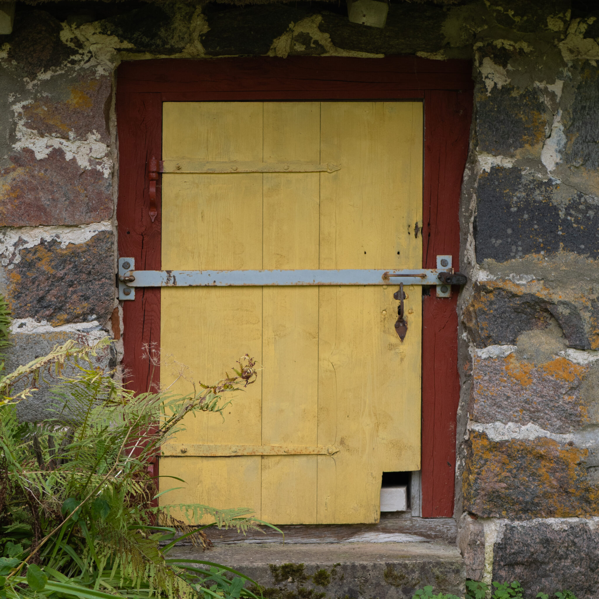 Kollane uks punase raamiga ja lukuga kiviseinas.