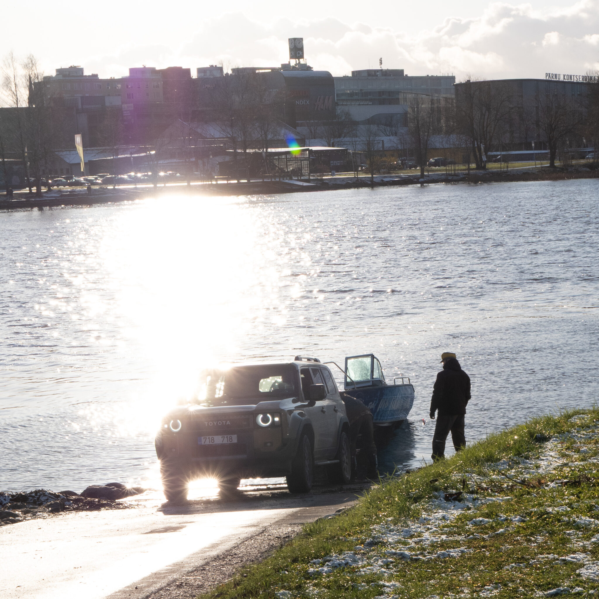 Toyota linnamaastur järve ääres, paat vees ja linn taustal.