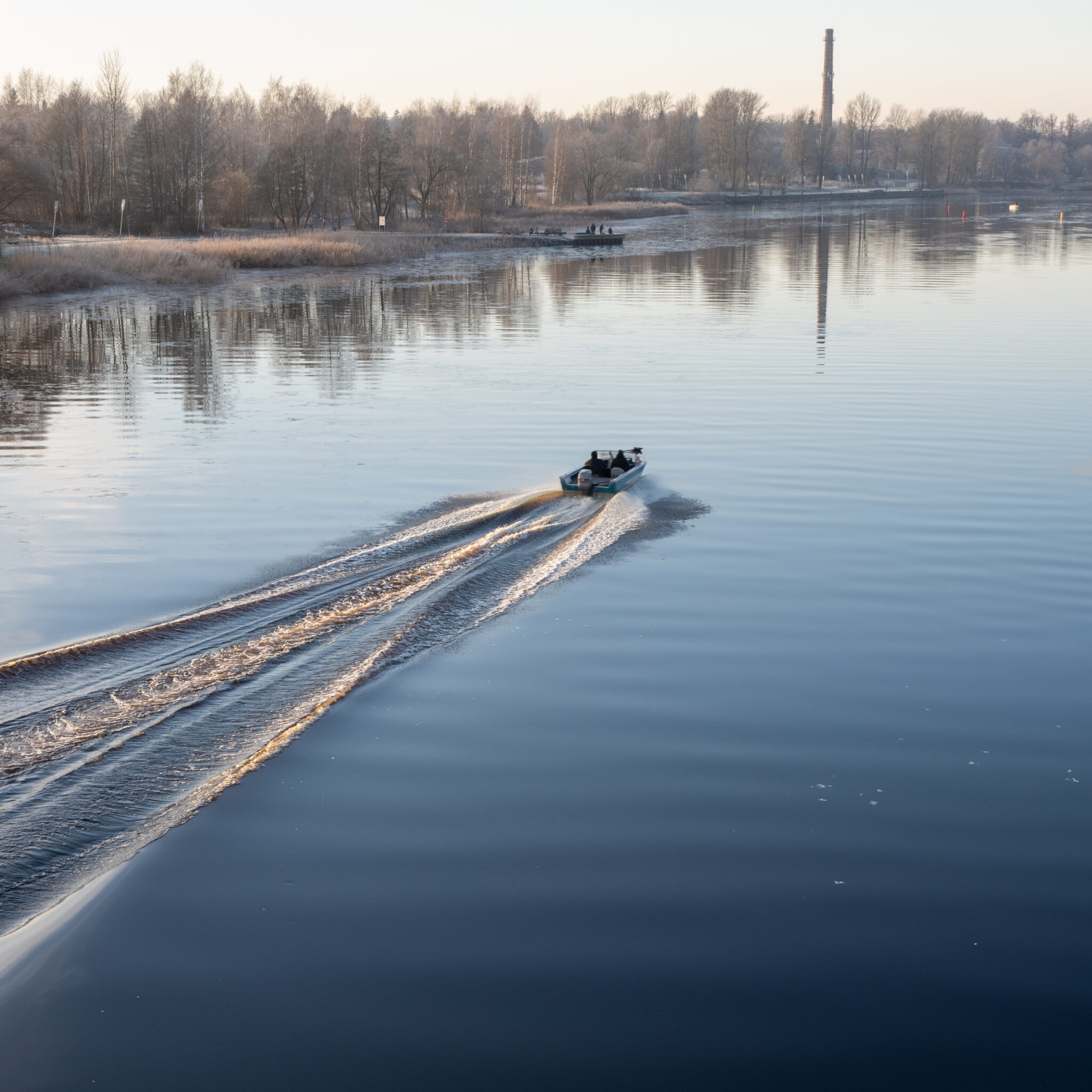 Paat jätab rahulikule järvele jäljevahu, kauguses tööstuskorsten.