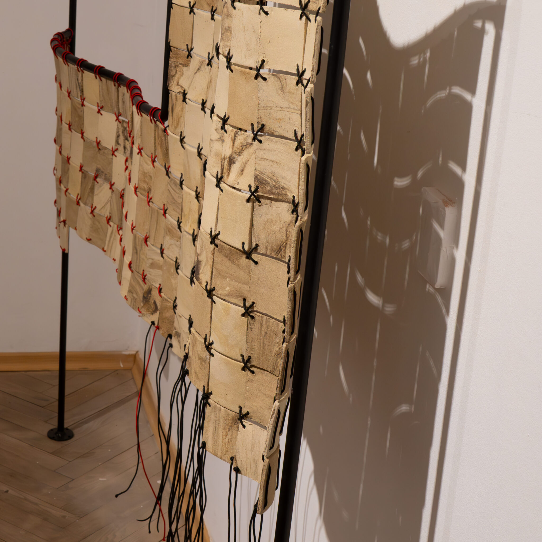 Punaste ja mustade niitidega tekstiiliteos ripub posti küljes seina ees.