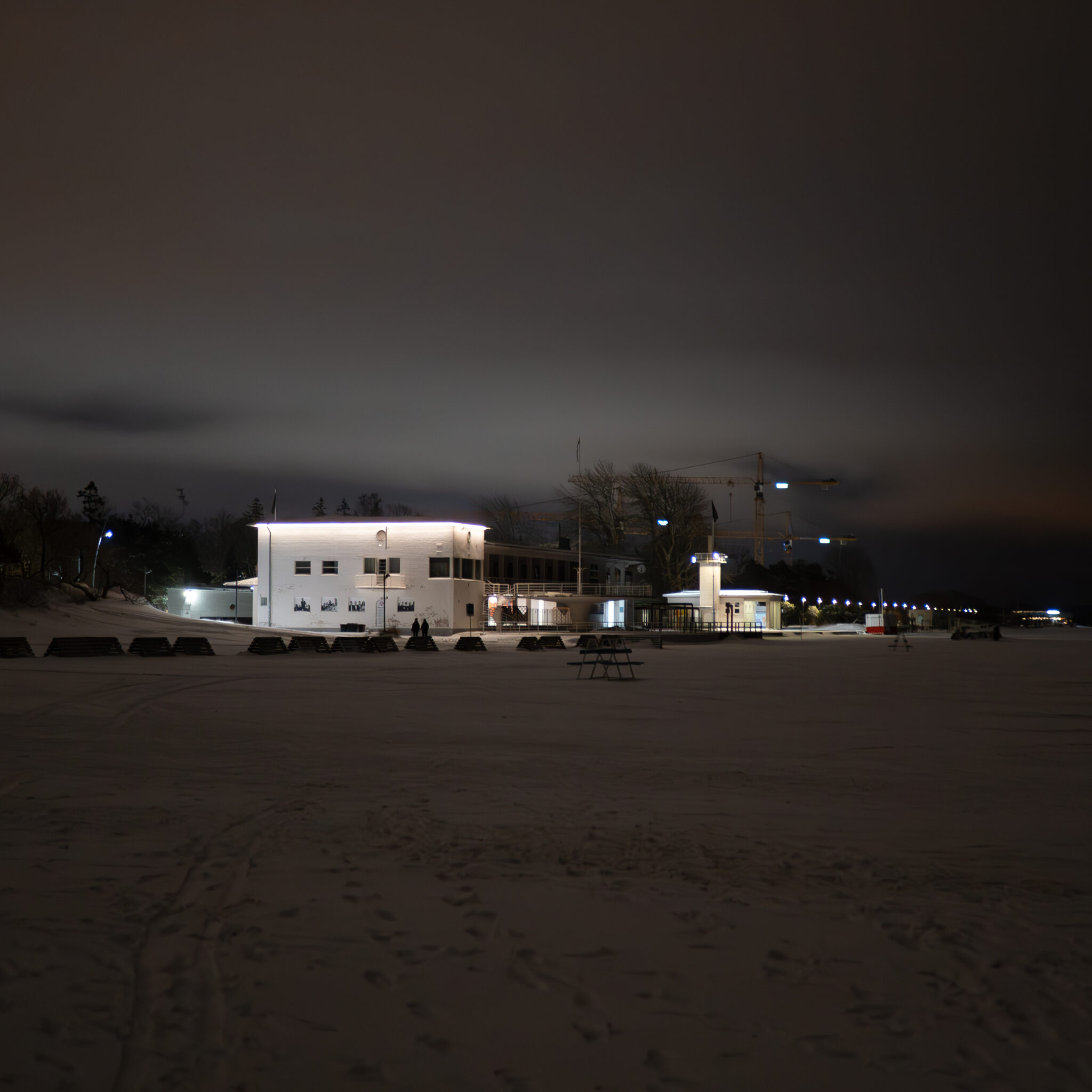 Öine rand valge hoone ja kauguses paistvate kraanadega.