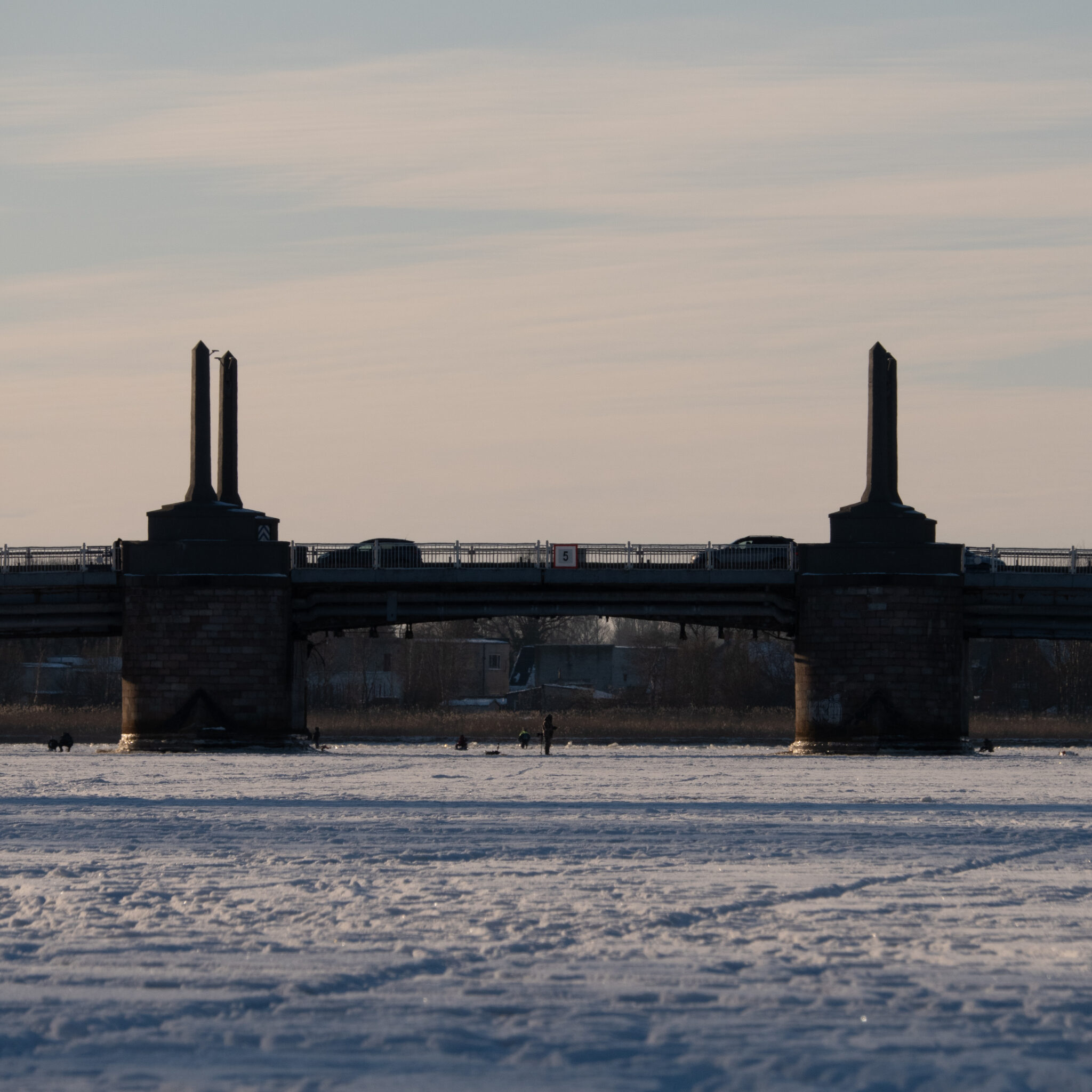 Sild ulatub üle jäätunud jõe, taustal sambad ja mõned inimesed.